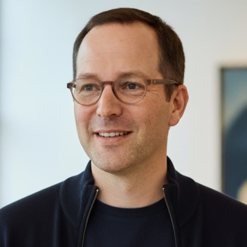 Smiling man with brown hair, wearing glasses and a dark blue zippered jacket.