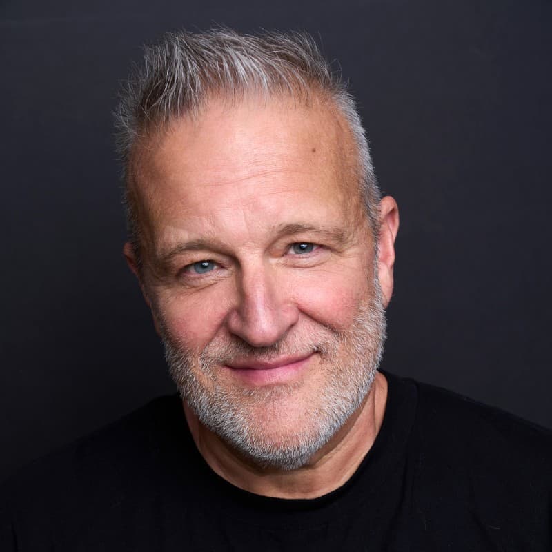 Friendly man with blue eyes, gray hair, and beard smiling against a dark background.