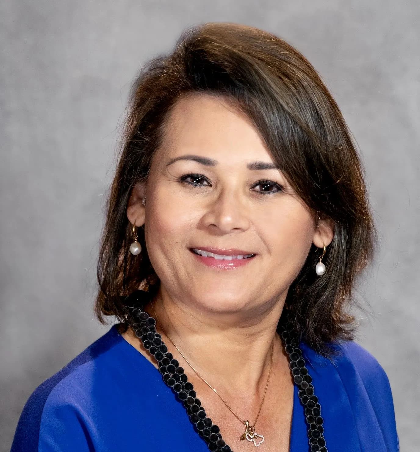 Smiling woman with short brown hair, blue top, black necklace, and pearl earrings.